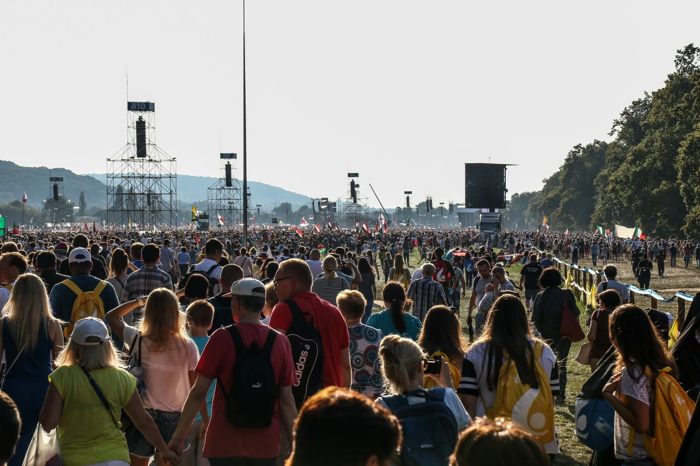 Młodzi wierni podczas Światowych Dni Młodzieży tłum ludzi zgromadzony na otwartym terenie podczas festiwalu lub dużego zgromadzenia, widoczne barierki oddzielające uczestników, rozstawione maszty techniczne oraz duża scena w oddali, po bokach widać drzewa, wiele osób niesie plecaki i flagi