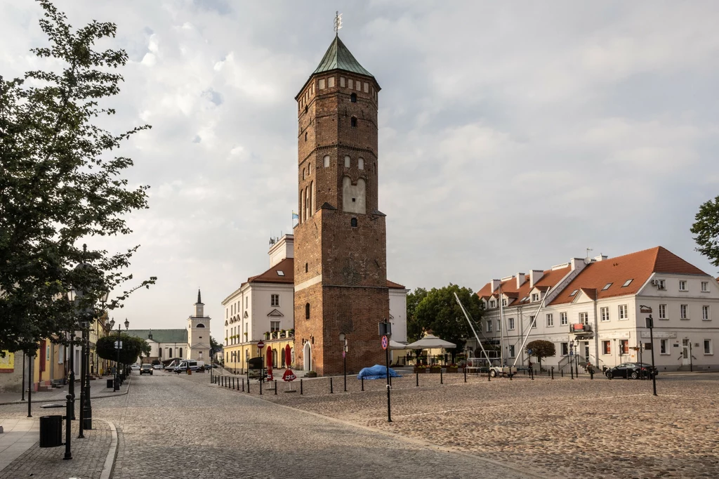 Autorka bloga "Mikrowyprawy z Warszawy" zachęca do odwiedzenia Pułtuska Kamienna wieża o charakterze historycznym dominuje nad brukowanym placem rynku, wokół którego stoją odrestaurowane, jasne kamienice z czerwonymi dachami; w tle widoczny jest niewielki kościół oraz pojedyncze zaparkowane samochody.