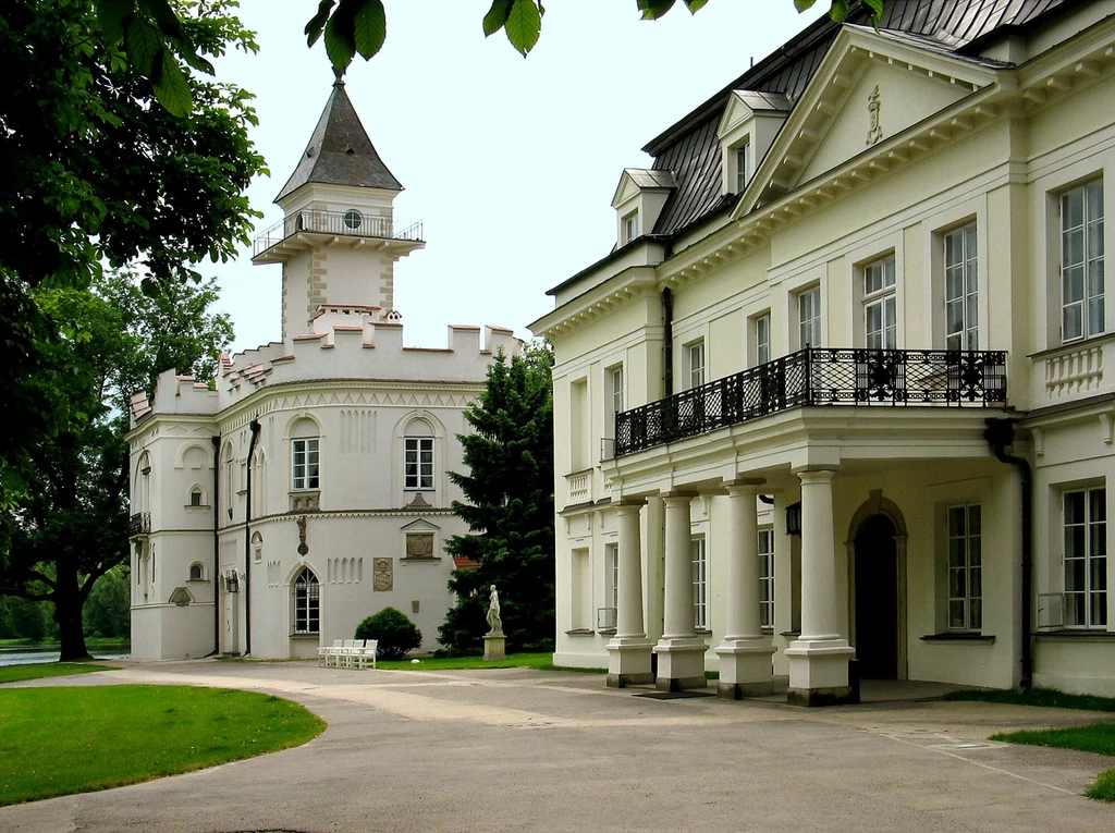Pałac w Radziejowicach Historyczny pałac z wieżą, otoczony zielenią, z eleganckimi białymi fasadami i ozdobnym balkonem, na spokojnym, zadbanym terenie z alejką oraz starodrzewem wokół.