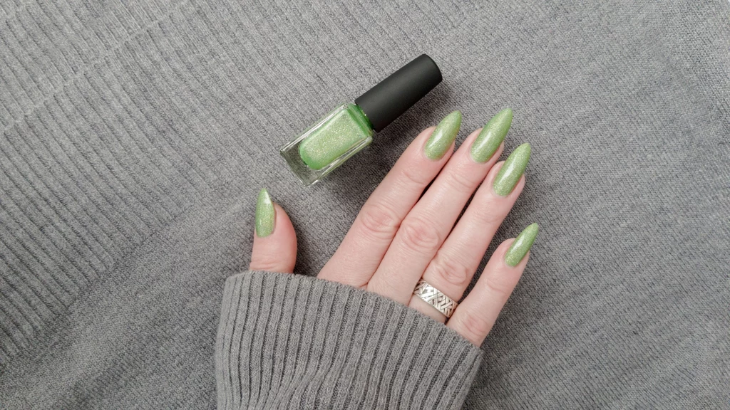 Trendy w manicure na jesień 2025 Dłoń z długimi paznokciami pomalowanymi zielonym lakierem oparta o szare, prążkowane ubranie, obok znajduje się buteleczka zielonego lakieru do paznokci.