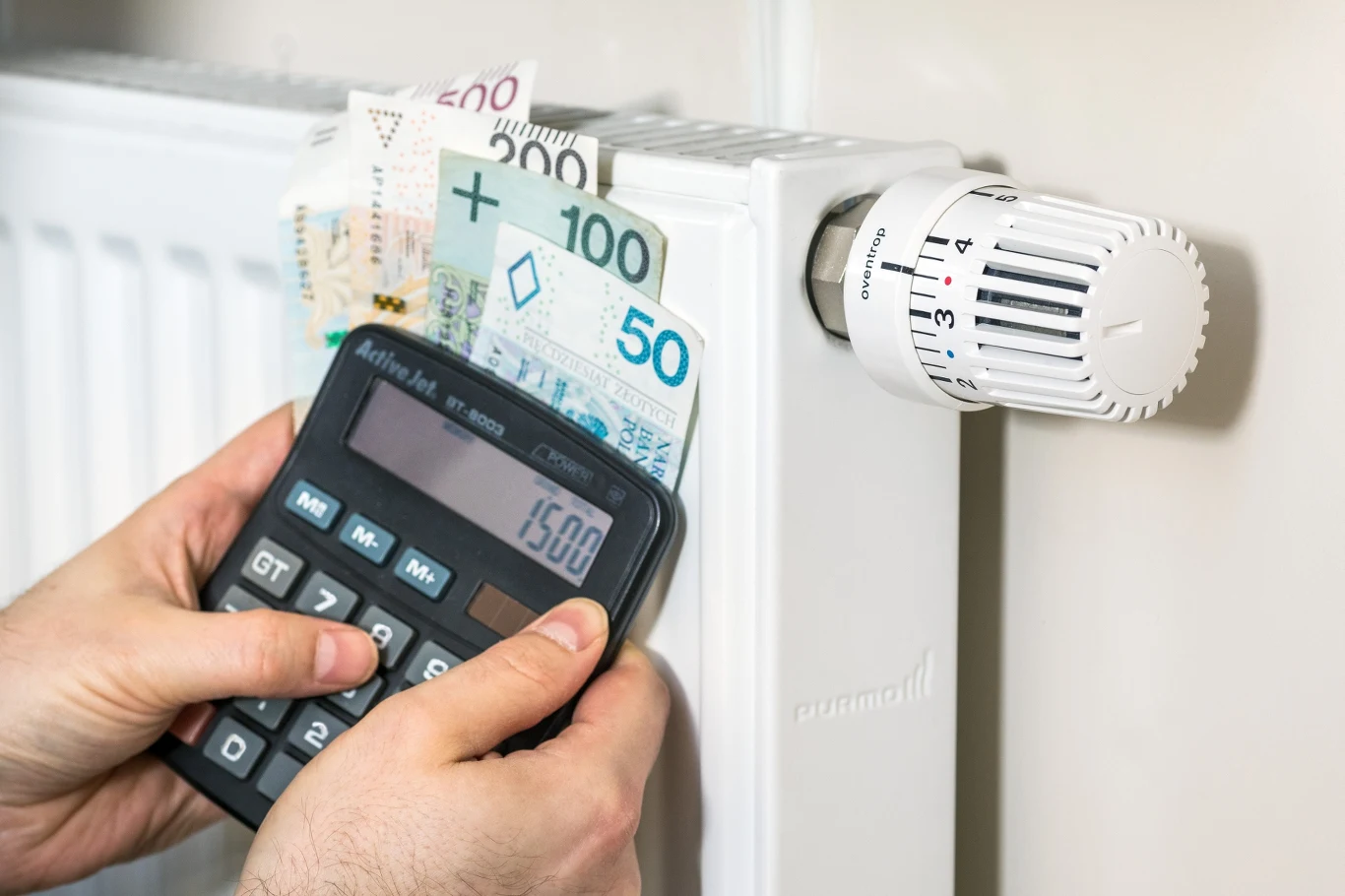 W nadchodzącym sezonie wzrosną rachunki za ogrzewanie. Rząd szykuje wsparcie dla najuboższych rodzin Ręce trzymające kalkulator oraz polskie banknoty przy kaloryferze z ustawionym termostatem, co akcentuje problem wyliczania kosztów ogrzewania.