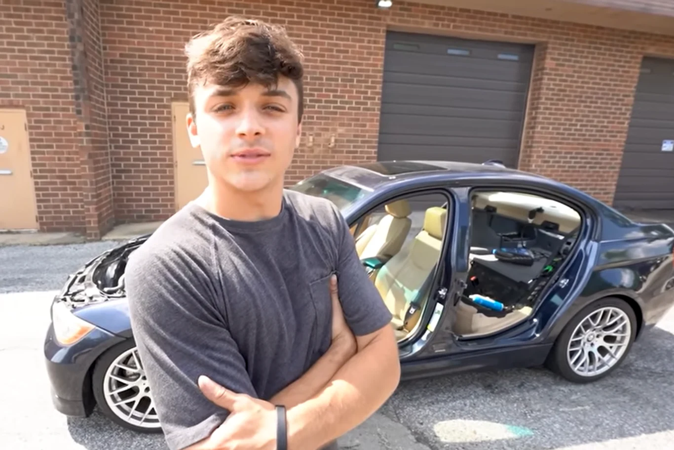 Młody mężczyzna stoi z założonymi rękami na tle czarnego samochodu z otwartymi drzwiami i podniesioną maską, na parkingu obok budynku z cegły.