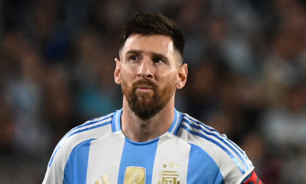 Lionel Messi Mężczyzna z brodą w stroju piłkarskim Argentyny, skupiony, z opaską kapitana na ramieniu, na tle rozmytego stadionu.