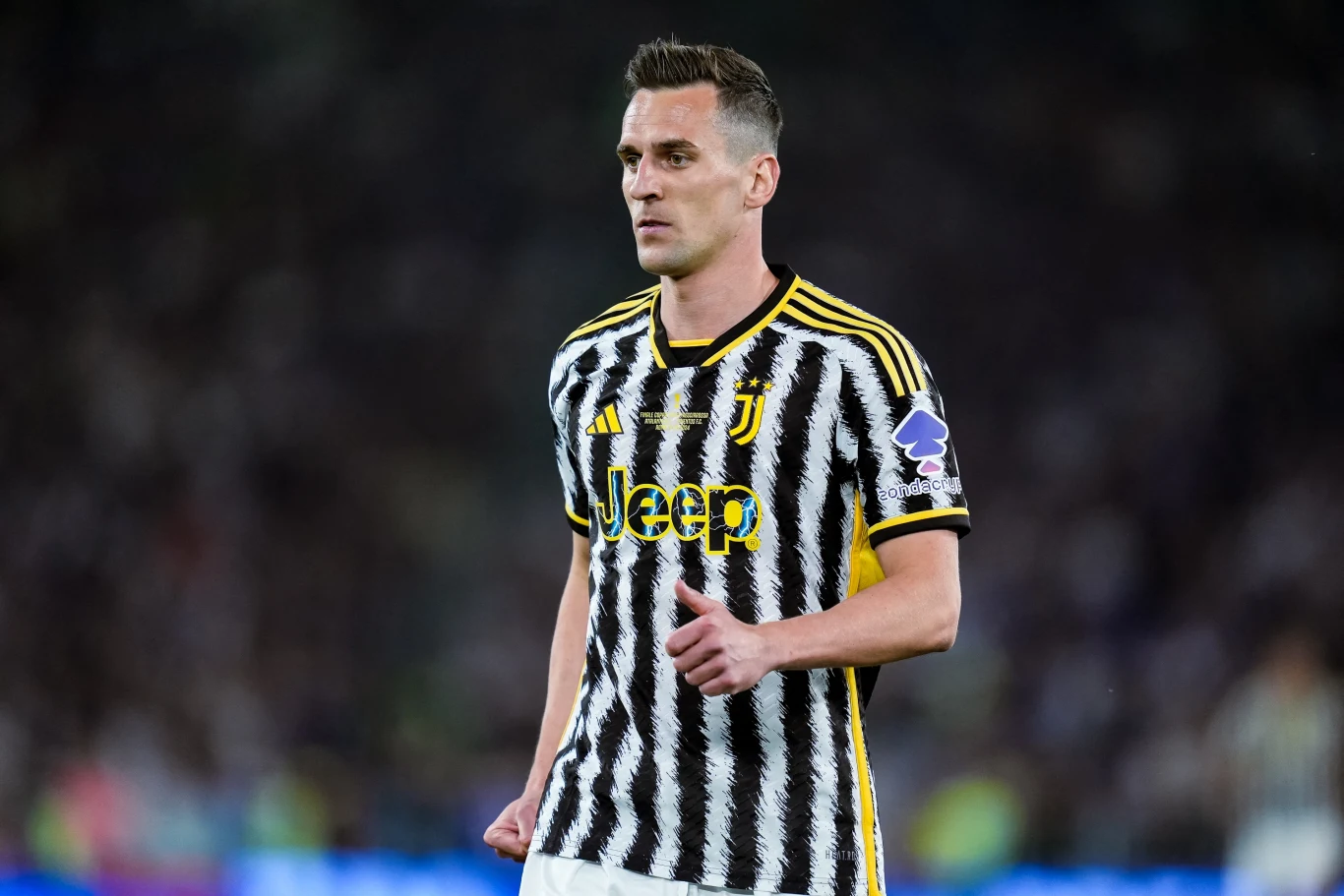 Piłkarz w biało-czarnej koszulce z żółtymi akcentami podczas meczu, na tle rozmytego stadionu wypełnionego kibicami.