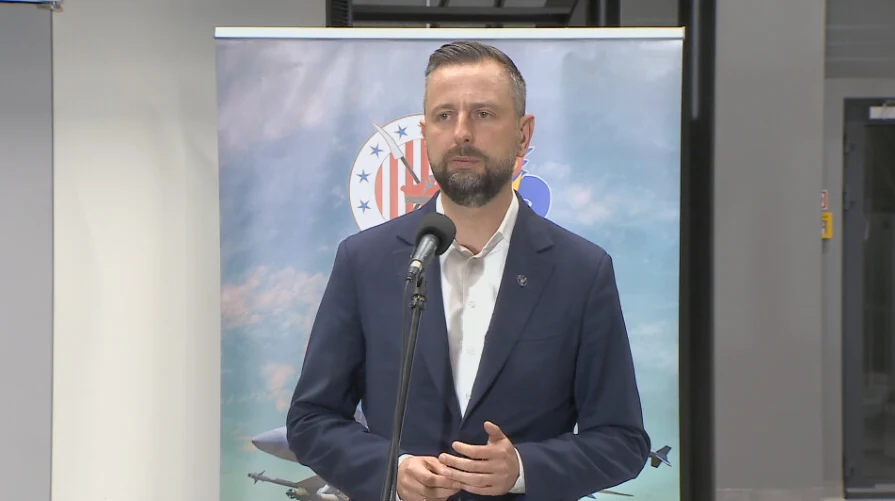 Mężczyzna w garniturze stoi przed mikrofonem na tle plakatu z symbolami wojskowymi oraz samolotami.