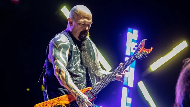 Muzyk z tatuażami na rękach grający na charakterystycznej, jaskrawo pomarańczowej gitarze elektrycznej na tle kolorowych świateł scenicznych, dynamiczna atmosfera koncertu.