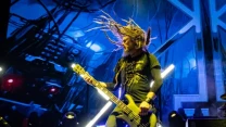 Muzyk z dredami grający na gitarze basowej na scenie koncertowej, dynamiczne oświetlenie i futurystyczna scenografia w tle.