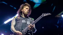 Gitarzysta na scenie podczas koncertu gra na elektrycznej gitarze typu V, ma rozpuszczone włosy, widoczne są tatuaże na ramieniu oraz kamizelka z naszywkami, w tle widać oświetlenie sceniczne.