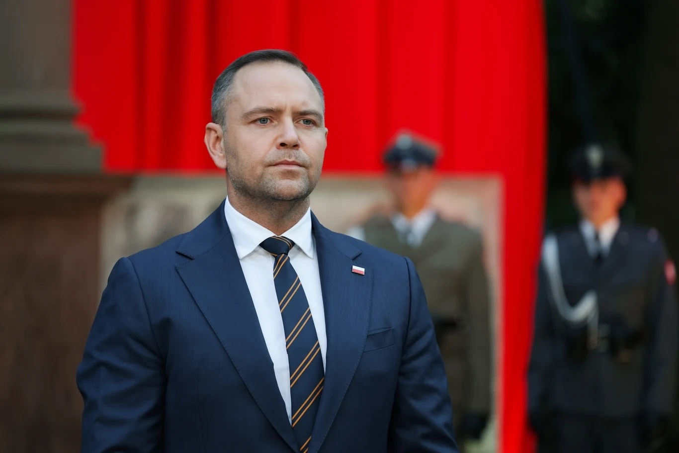 Mężczyzna w garniturze z przypiętą flagą Polski stoi na tle czerwonej kurtyny, w dalszym planie widoczni dwaj mundurowi strażnicy w wojskowych i policyjnych mundurach.