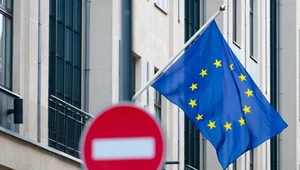 Flaga Unii Europejskiej zawieszona na budynku w jasnym kolorze. Na pierwszym planie rozmazany znak zakazu, wyostrzenie obejmuje wszystko za nim, ale nie jego.