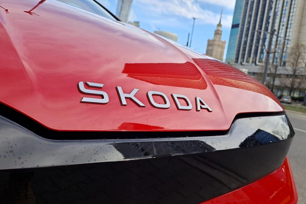 Z tyłu i przodu Elroqa nie znajdziemy już charakterystycznego logo czeskiej marki. Zastąpił je napis "SKODA". Czerwony samochód marki Skoda na tle nowoczesnych wieżowców i Pałacu Kultury i Nauki w centrum miasta, widoczny z bliska przód pojazdu z wyraźnym logo producenta.