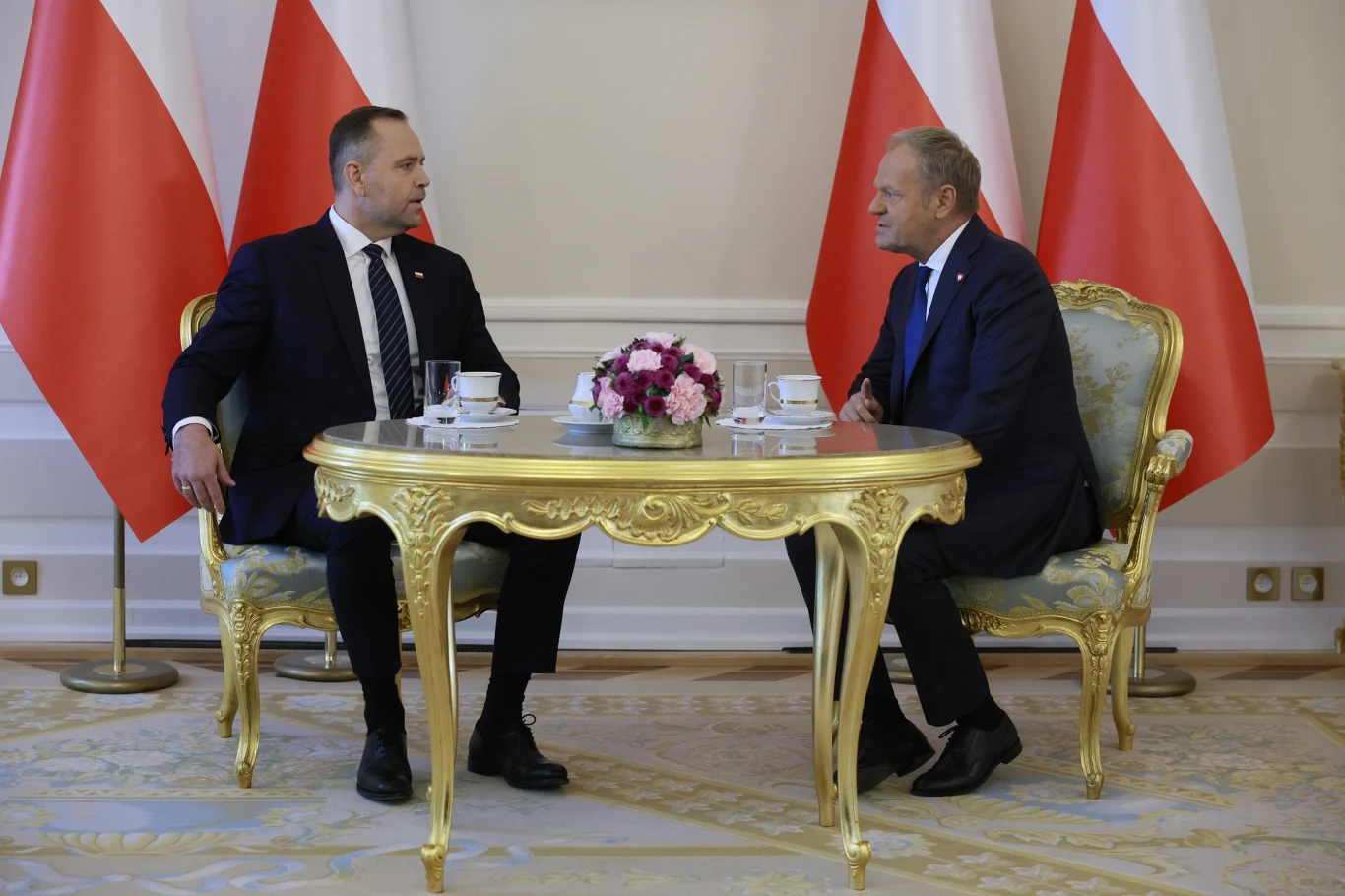 Współpraca Tuska z Nawrockim zaczyna się łagodnie Dwóch mężczyzn w garniturach siedzi naprzeciw siebie przy ozdobnym, złotym stole z kwiatami, w tle widoczne biało-czerwone flagi.