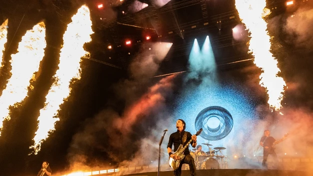 Zespół rockowy grający na dużej scenie w otoczeniu efektów pirotechnicznych z płomieniami i światłami, tło wypełniają dymy i dynamiczna oprawa wizualna, atmosfera żywiołowego koncertu.