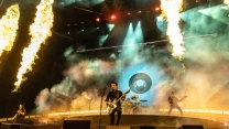 Zespół muzyczny występuje na dużej scenie z efektami pirotechnicznymi, kolorowym oświetleniem i dymem, na pierwszym planie wokalista z gitarą, w tle perkusista, dynamiczna atmosfera koncertu.