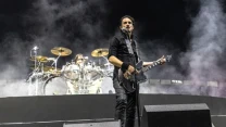 Gitarzysta i wokalista rockowy występuje na scenie z mikrofonem, z tyłu widoczny perkusista oraz efektowna oprawa świetlna i dym sceniczny, scena otoczona dużymi głośnikami.