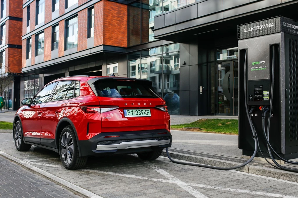 Skoda Elroq na jednym ładowaniu może przejechać 573 km. Czerwony samochód elektryczny marki Škoda podłączony do publicznej stacji ładowania przy nowoczesnym budynku biurowym, kabel ładowarki podłączony do samochodu, asfaltowa droga i chodnik w otoczeniu, szklana elewacja budynku w tle.