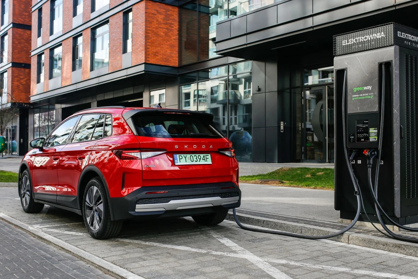 Czerwony samochód elektryczny marki Škoda podłączony do publicznej stacji ładowania przy nowoczesnym budynku biurowym, kabel ładowarki podłączony do samochodu, asfaltowa droga i chodnik w otoczeniu, szklana elewacja budynku w tle.