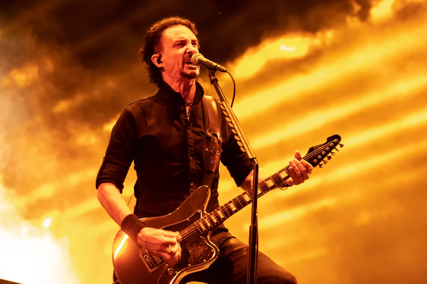 Joe Duplantier (Gojira) Energetyczny muzyk grający na gitarze elektrycznej i śpiewający do mikrofonu, dynamiczne światła w ciepłych barwach tworzą efektowne tło koncertowe.