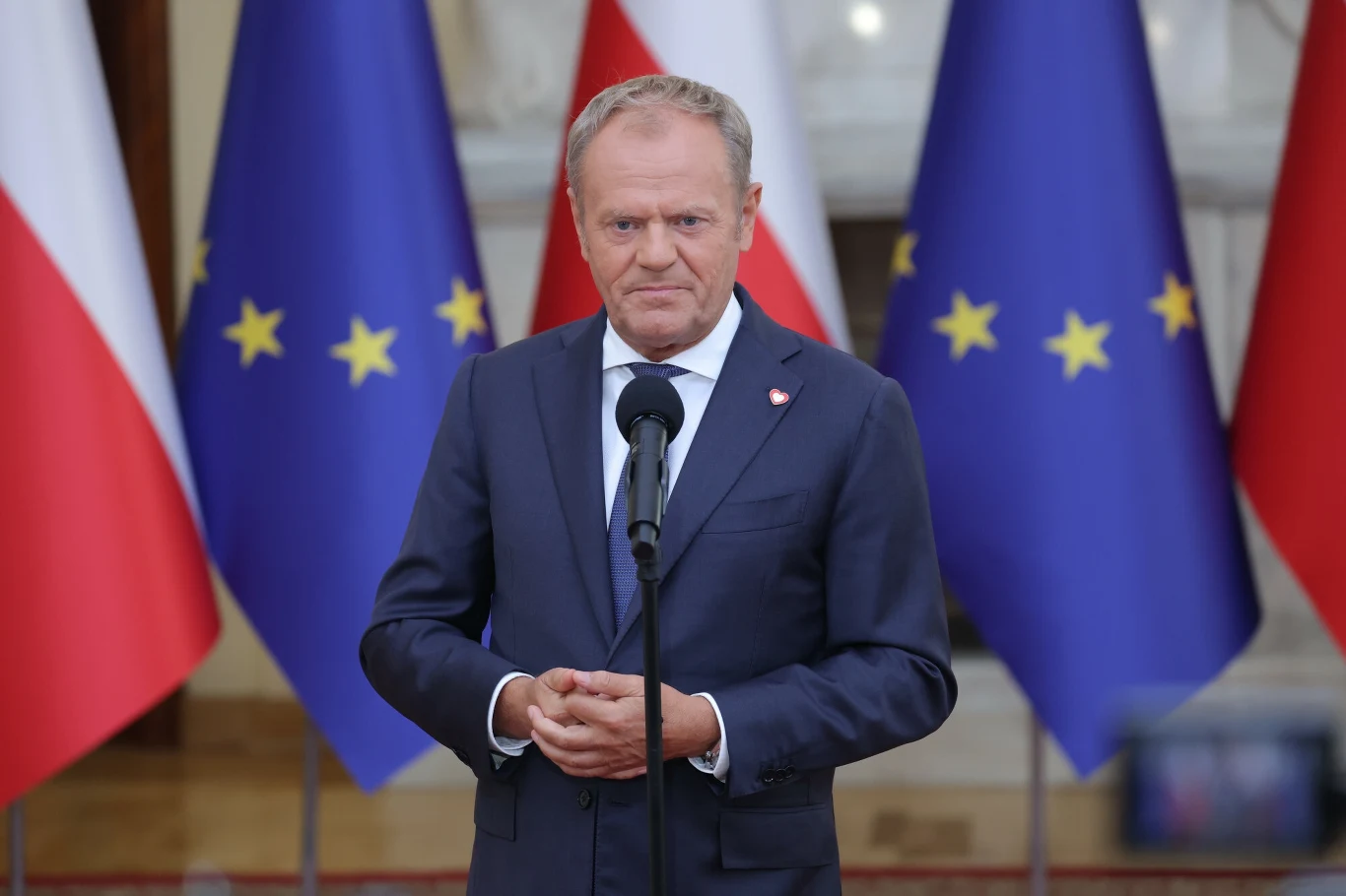 Premier Donald Tusk Mężczyzna w garniturze stoi przy mikrofonie, za nim znajdują się flagi Polski oraz Unii Europejskiej.