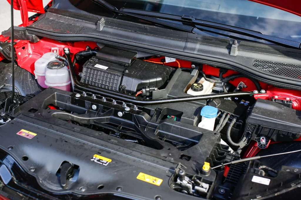 W standardzie Skoda Elroq oferuje 170 KM i napęd na tylne koła. Komora silnika samochodowego z widocznymi podzespołami, takimi jak filtr powietrza, zbiorniczki z płynami technicznymi oraz przewody i elementy mechaniczne umieszczone w czerwonym nadwoziu.