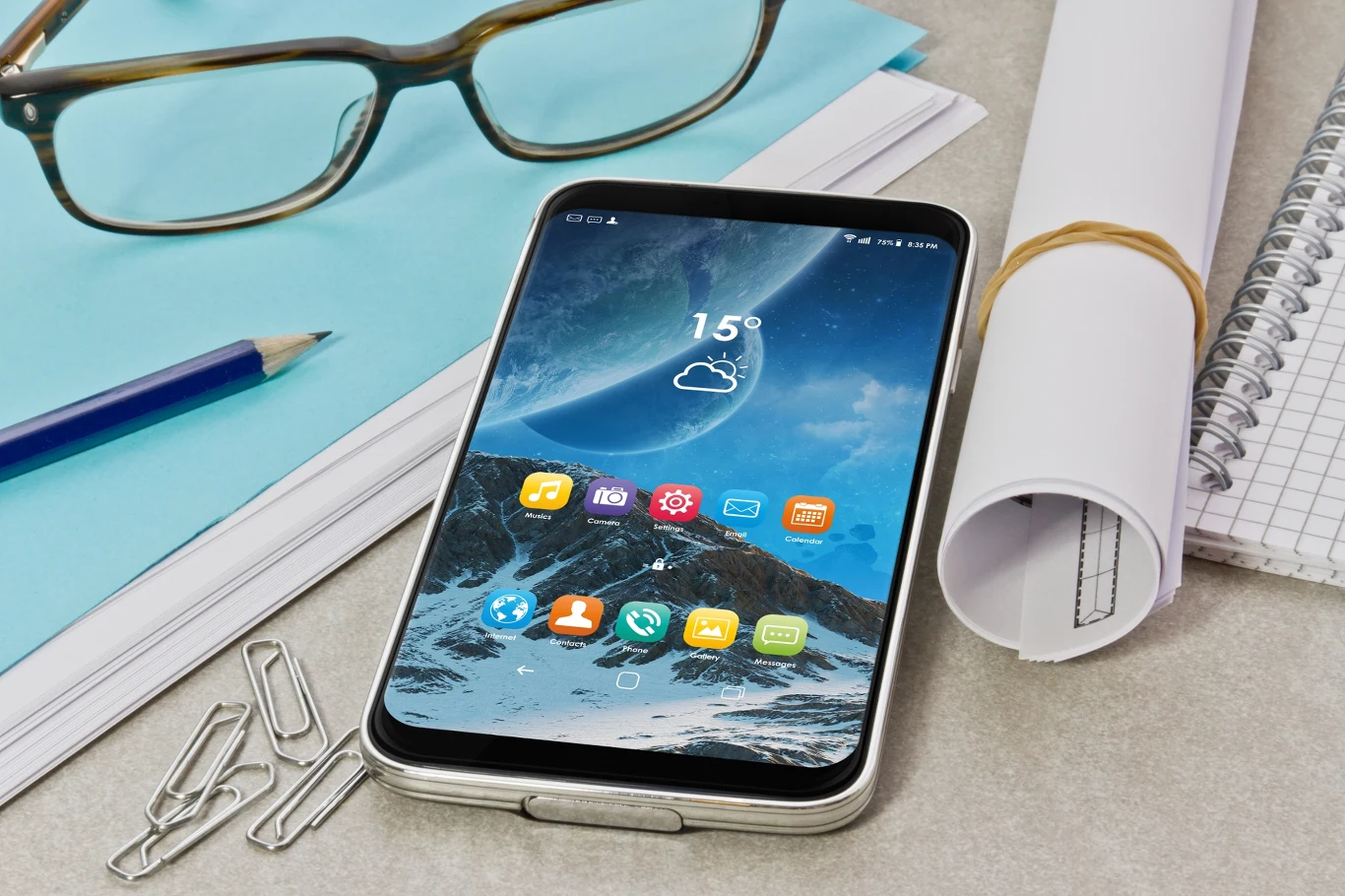 Nie chcesz, aby ktoś widział twoją aplikację bankową? Możesz ukryć ją z widoku. Smartfon leżący na biurku obok okularów, spinaczy, zrolowanego arkusza papieru, notesu w kratkę i długopisu, na ekranie wyświetlana jest aplikacja pogodowa i kolorowe ikony.