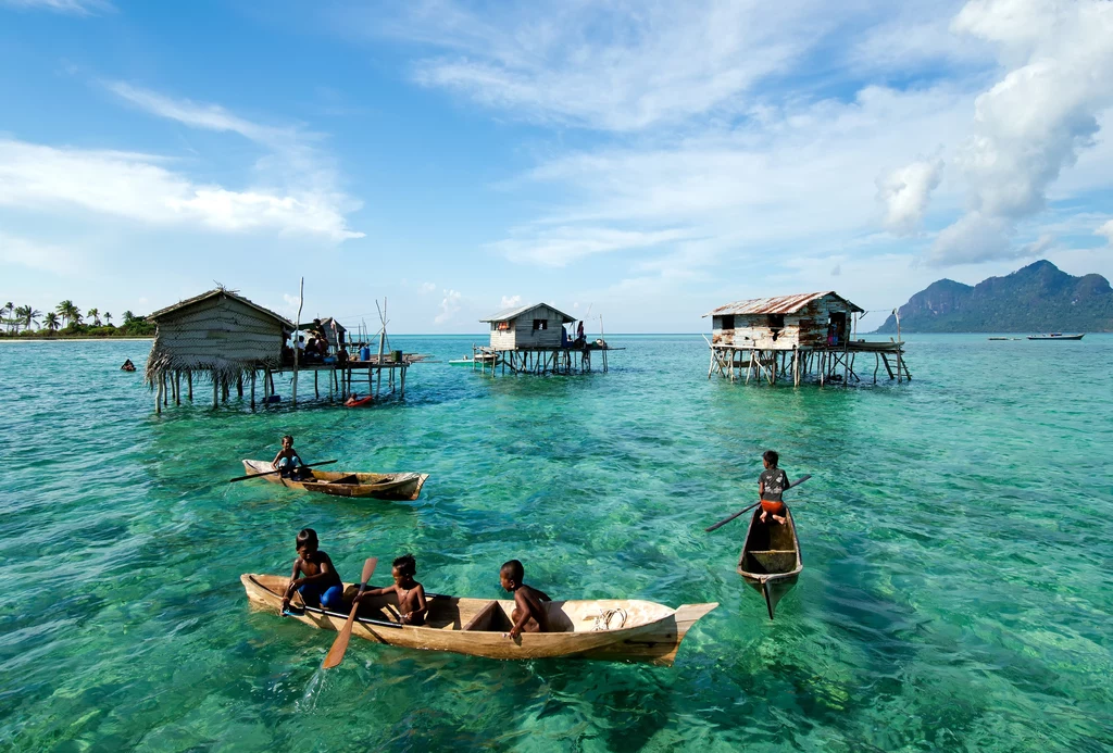 Bajau - morscy nomadzi Drewniane chaty na palach wznoszą się nad przezroczystą, turkusową wodą, po której dzieci w kilku drewnianych łódkach pływają na tle tropikalnego krajobrazu i gór widocznych w oddali.