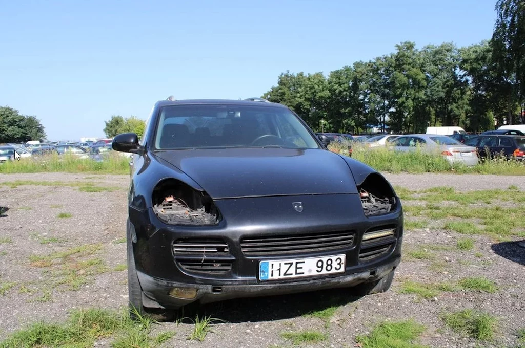 Czarny samochód marki Porsche Cayenne z brakującymi reflektorami, widoczny od frontu na parkingu otoczonym przez inne zaparkowane pojazdy oraz drzewa w tle.