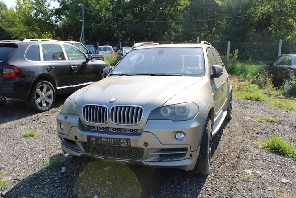 SUV marki BMW w kolorze jasnym stoi zaparkowany na żwirowym placu, obok innego samochodu tej samej marki. W tle widoczne drzewa, ogrodzenie z siatki oraz inne zaparkowane pojazdy.