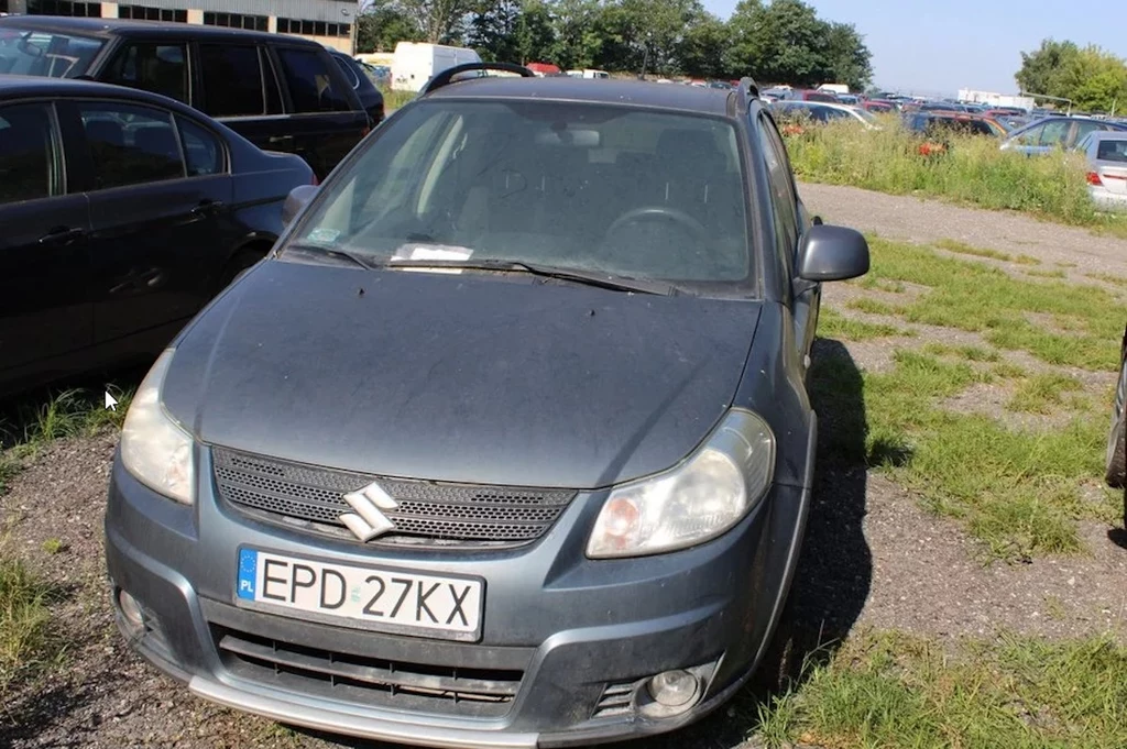 Szary samochód osobowy marki Suzuki stoi zaparkowany na nieutwardzonym parkingu, otoczony innymi pojazdami oraz trawą. Na przedniej szybie widoczny jest dokument, prawdopodobnie mandat lub informacja parkingowa. Auto jest lekko zakurzone, a tablica rej...