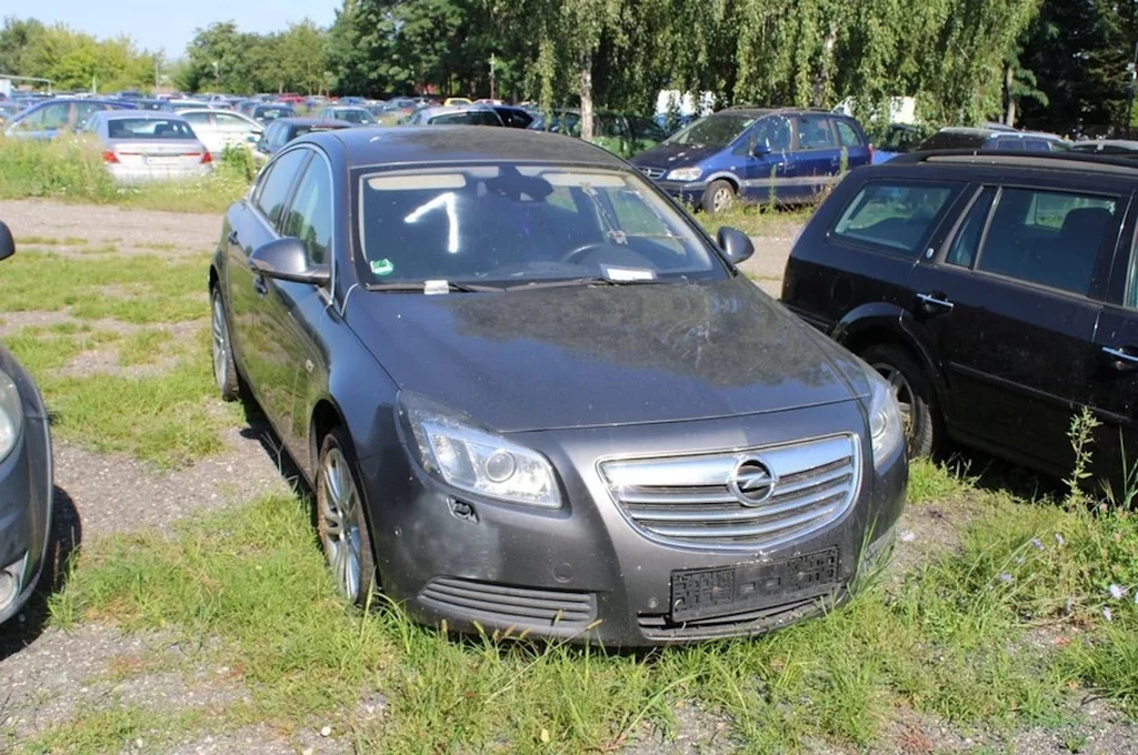 Czarny samochód marki Opel zaparkowany na nieutwardzonym parkingu, widoczne ślady zabrudzenia i braku tablic rejestracyjnych, w tle inne pojazdy oraz zieleń i drzewa.