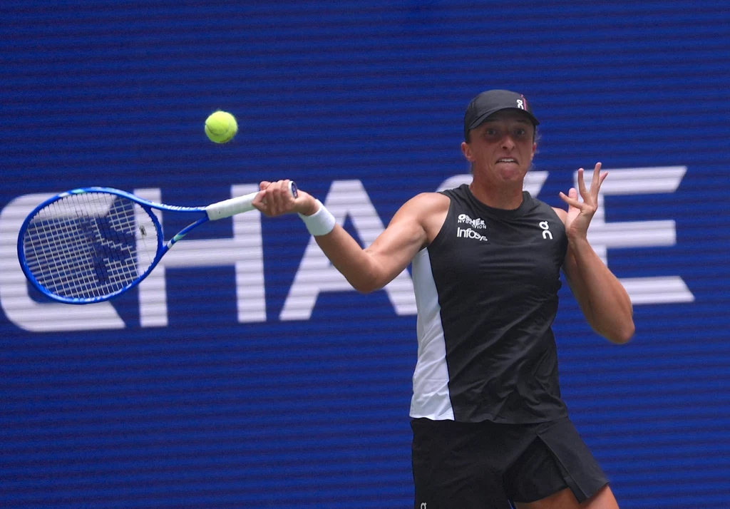 Iga Świątek podczas turnieju miksta US Open 2025 Tenisistka w czarnej koszulce i czapce uderza piłkę rakietą podczas meczu, dynamiczna poza i skoncentrowany wyraz twarzy, w tle niebieska ściana z widocznym fragmentem napisu.