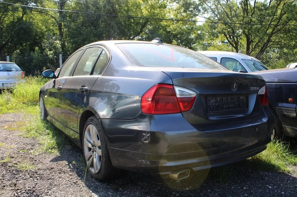 Szary sedan marki BMW zaparkowany na trawiastym parkingu obok innych pojazdów, światło słoneczne tworzy odblask na tylnej szybie, wokół auta rośnie wysoka trawa, w tle drzewa i ogrodzenie z siatki.