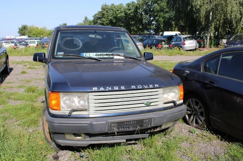 Stary ciemnoniebieski Range Rover zaparkowany na trawiastym parkingu, widoczne ślady zużycia na zderzaku i karoserii, częściowo zasłonięta tablica rejestracyjna, w tle inne zaparkowane samochody oraz drzewa.
