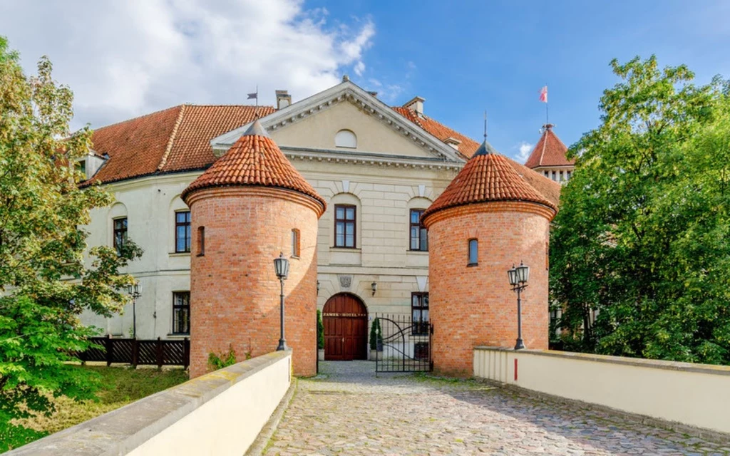 Pułtusk to jedno z najstarszych miast na Mazowszu Zabytkowy budynek z czerwonej cegły z dwiema cylindrycznymi wieżami przykrytymi stożkowatymi dachami, otoczony zielenią, kamiennym mostkiem i klasyczną fasadą z drewnianymi drzwiami pośrodku.