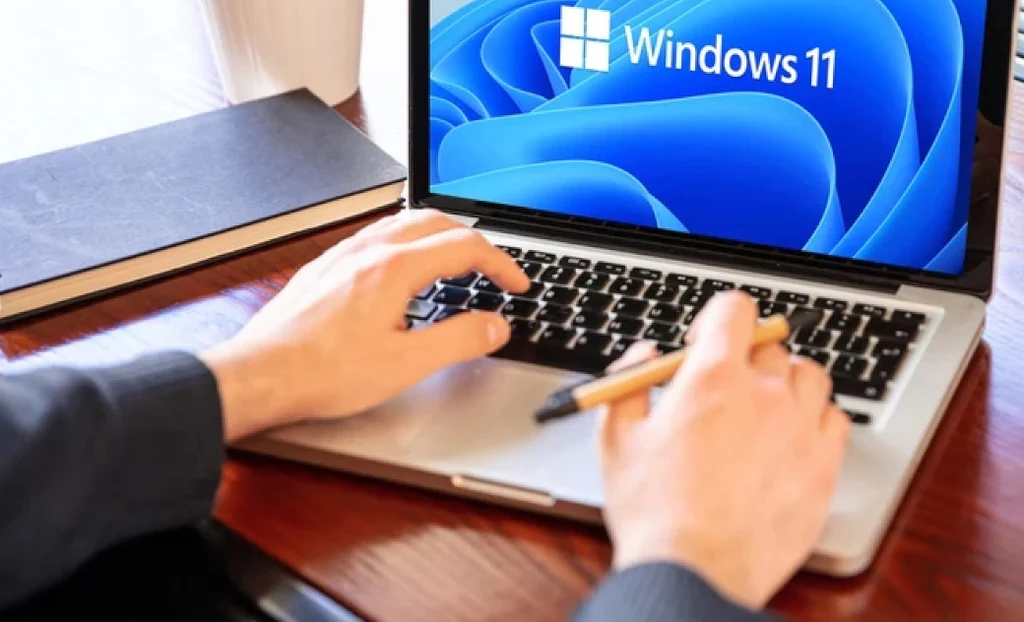 Czy nowa odsłona popularnego komunikatu o błędzie Windowsa spotka się z pozytywnym odzewem użytkowników? Osoba korzysta z laptopa, na którego ekranie wyświetla się ekran startowy systemu Windows 11, obok laptopa leży zamknięty czarny notes oraz długopis.