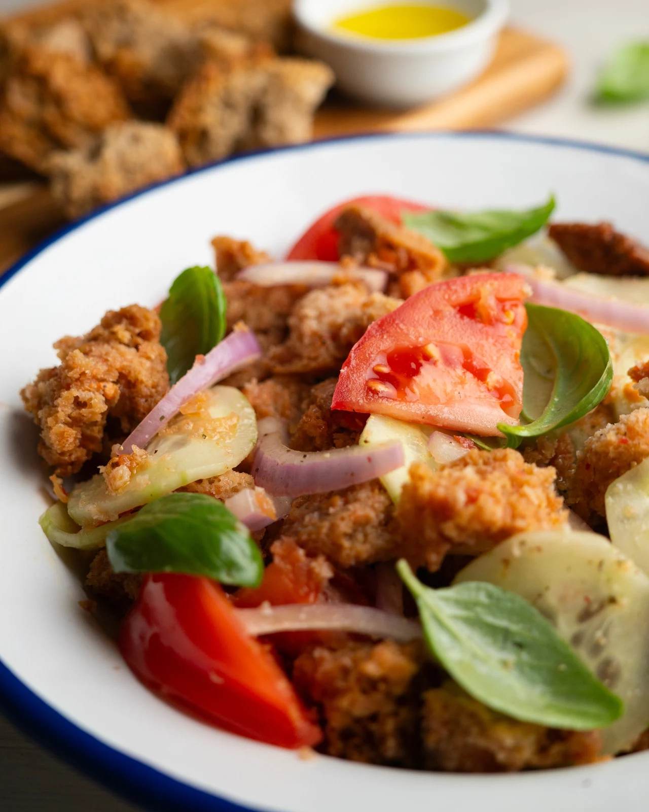 Panzanella to połączenie prostych składników. Smakuje wybornie! Sałatka z pokruszonym chlebem, plasterkami pomidorów, ogórków i czerwonej cebuli, udekorowana świeżymi liśćmi bazylii, podana w białej misce z niebieską obwódką; w tle kawałki chleba i miseczka z oliwą.