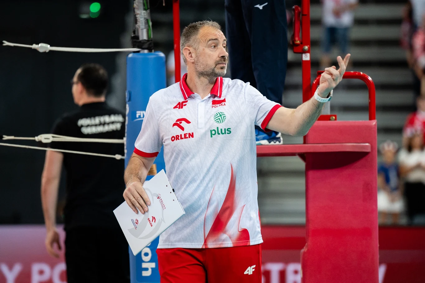 Trener siatkówki ubrany w biało-czerwony strój z logotypami sponsorów gestykuluje podczas meczu, trzymając notes w dłoni; w tle widoczne są fragmenty boiska oraz rozmyci kibice na trybunach.