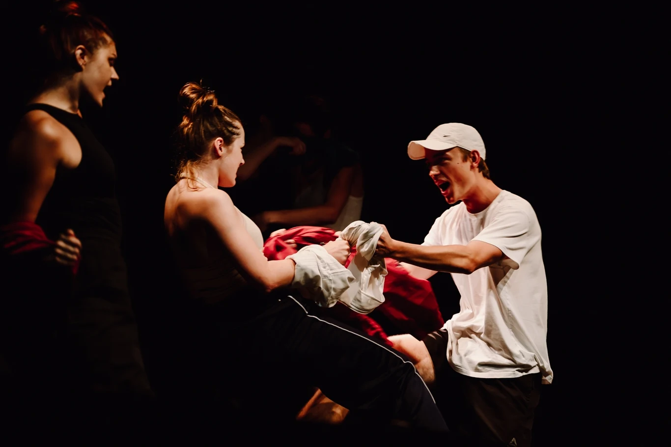 Trzy osoby w wyraźnie oświetlonym punkcie na scenie, jedna kobieta siedzi i ściska ubranie, mężczyzna w czapce krzyczy w jej kierunku, w tle widoczna postać w cieniu, atmosfera napięcia i ekspresji.