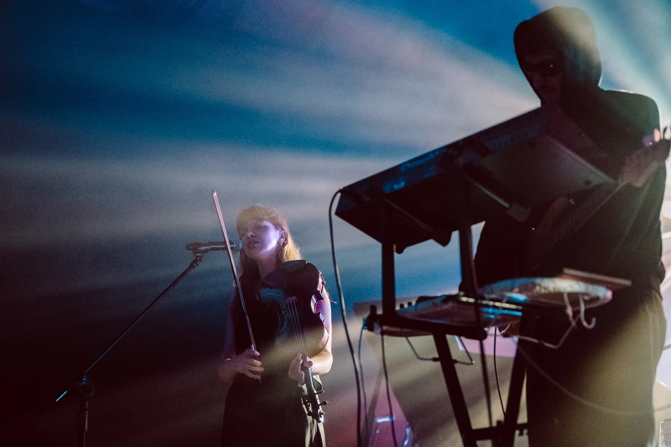 Dwóch muzyków na scenie, jeden gra na skrzypcach i śpiewa do mikrofonu, drugi obsługuje syntezator. Silne światła i dym tworzą klimatyczną aurę podczas koncertu.