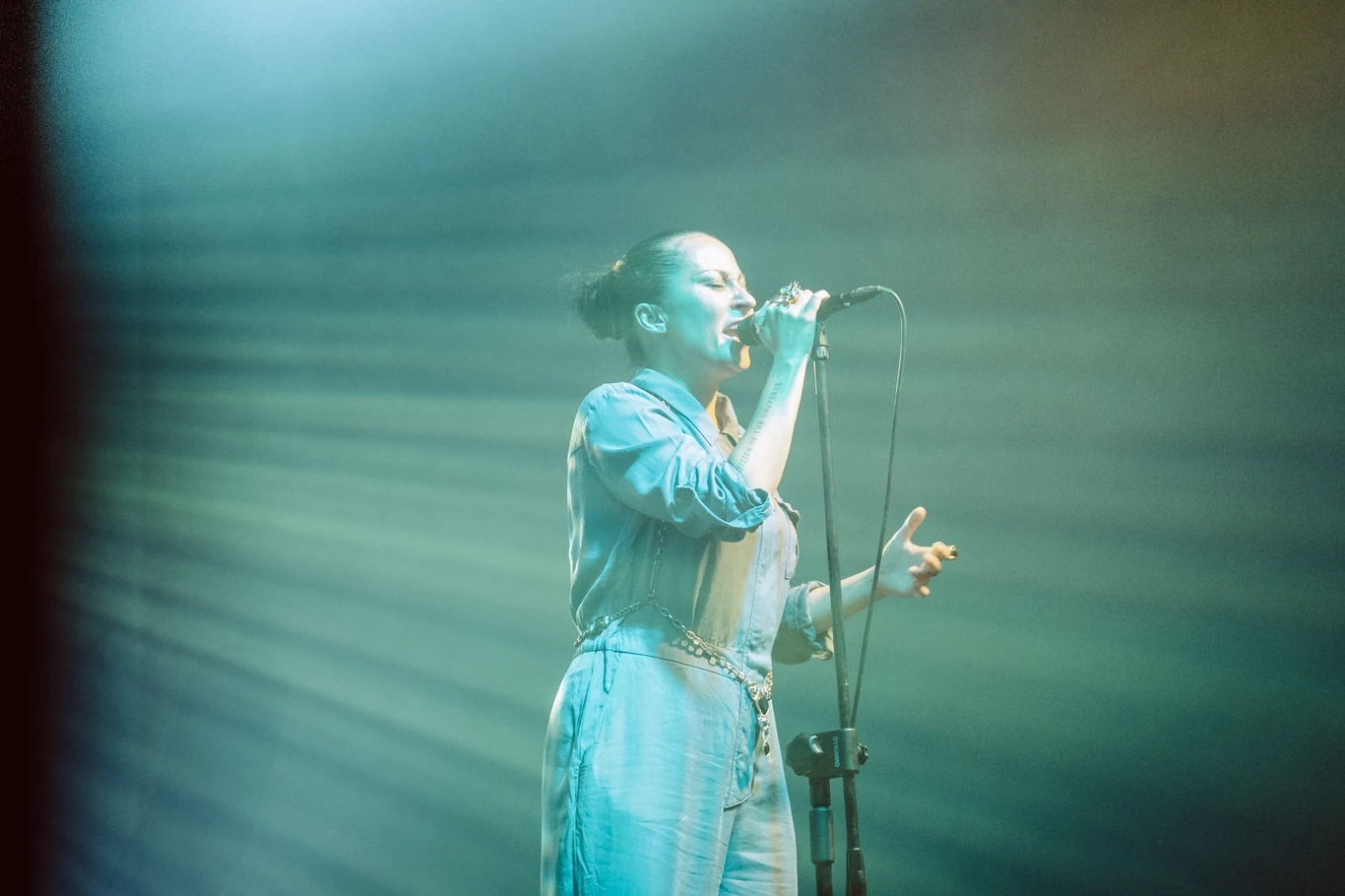 Kobieta w jasnym ubraniu śpiewa do mikrofonu na scenie, otoczona kolorowym, klimatycznym oświetleniem, z promieniami światła rozchodzącymi się w tle.