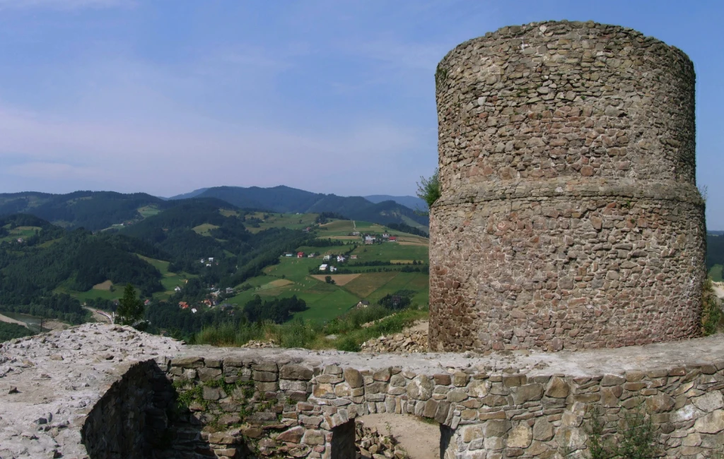 Zamek w Rytrze przechodzi metamorfozę. Pięknie położona budowla odzyskuje blask. Kamienna baszta i fragment murów obronnych ruin zamku na tle zielonych pagórków i rozległego krajobrazu wiejskiego, z widocznymi w oddali polami i małymi gospodarstwami.