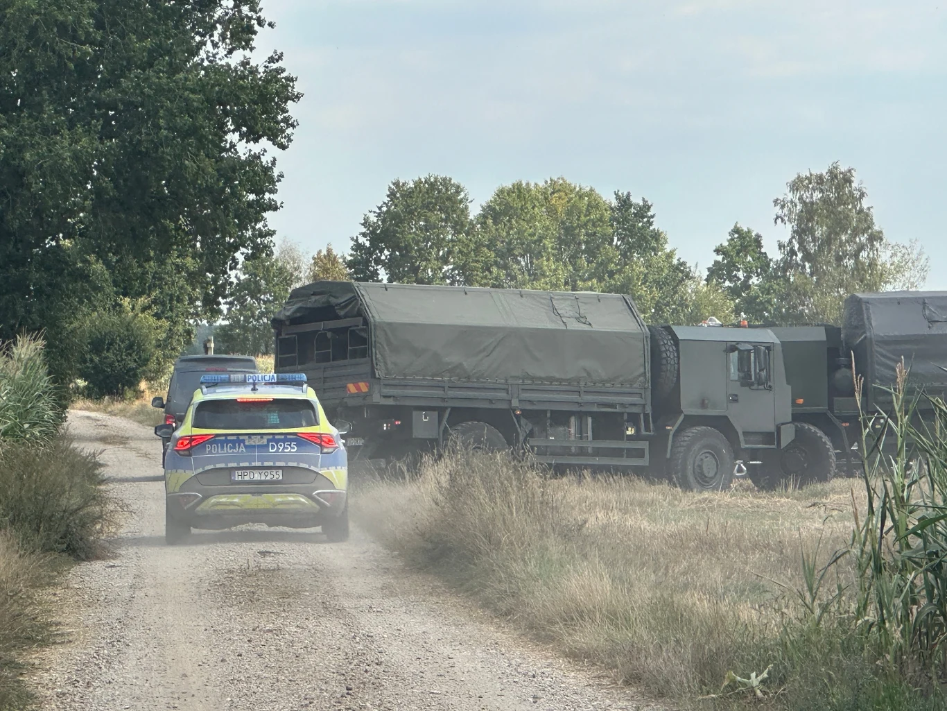 Na miejscu zdarzenia w Osinach pracuje policja i wojsko Radiowóz policyjny oraz wojskowe ciężarówki ustawione na polnej drodze, wokół teren porośnięty trawą i drzewami, atmosfera spokojna, bez obecności postaci ludzkich.