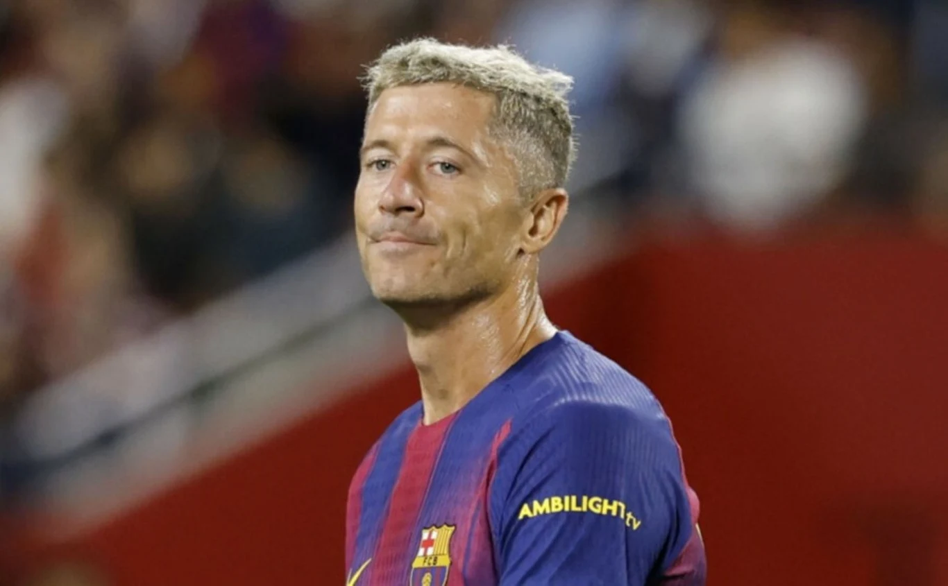 Robert Lewandowski Piłkarz w koszulce FC Barcelony stoi na boisku, patrząc w stronę widza, z widocznym wyrazem twarzy sugerującym zadumę lub rozczarowanie. Tło rozmazane z przewagą czerwieni i fioletu.