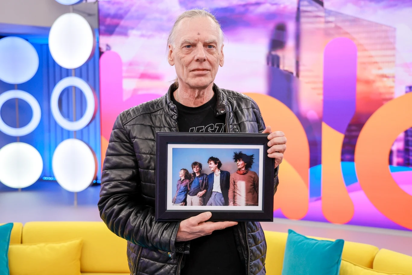 Tomek Lipiński 21 sierpnia kończy 70 lat Starszy mężczyzna w czarnej kurtce skórzanej stoi w nowoczesnym, kolorowym studiu telewizyjnym, trzymając w dłoniach oprawione zdjęcie grupy osób.