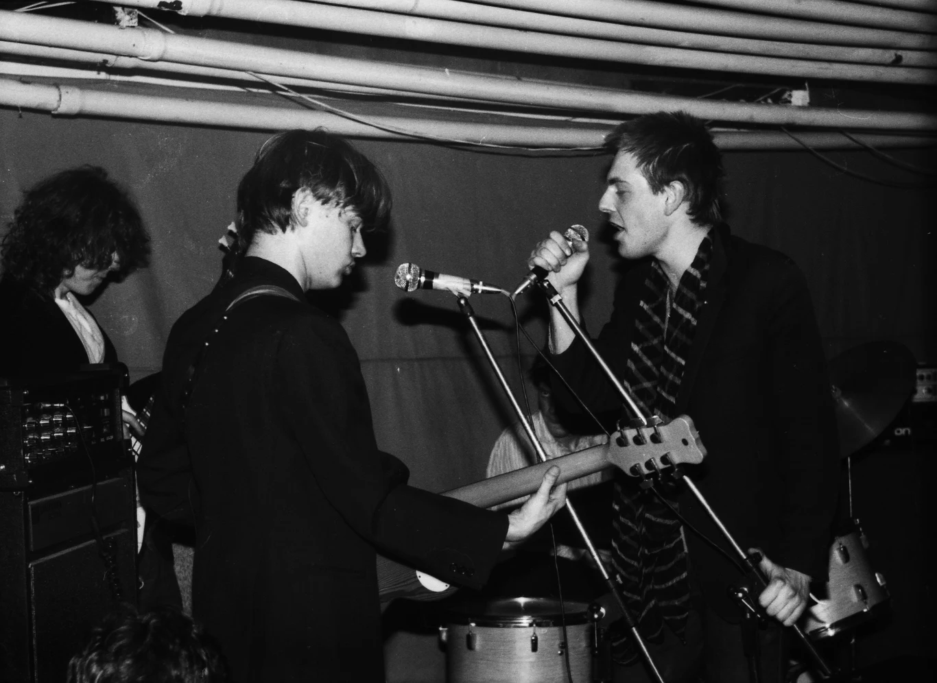 Robert Brylewski i Tomek Lipiński stworzyli Brygadę Kryzys Czterech muzyków podczas koncertu w kameralnej, podziemnej przestrzeni, jeden z nich śpiewa do mikrofonu, drugi gra na gitarze elektrycznej, widoczna perkusja w tle, całość utrzymana w czerni i bieli.