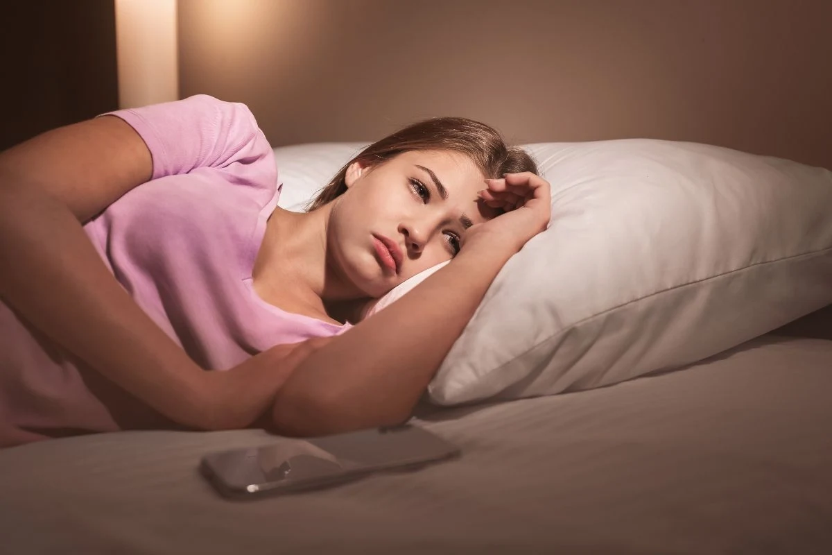 Młoda kobieta leżąca na łóżku z głową opartą o rękę, wyraźnie zamyślona lub zmartwiona, ubrana w różową koszulkę; obok na pościeli leży smartfon, oświetlenie w pokoju jest przytulne.