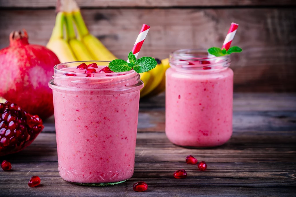 Warto dorzucić pestki granatu do smoothie lub wlać trochę naturalnego soku z tych owoców Dwa słoiki wypełnione różowym koktajlem z ziarnami granatu i listkami mięty, w tle leżą banany i owoc granatu, całość ustawiona na drewnianym blacie.