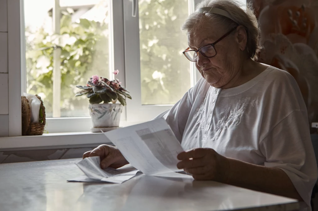 Starsza kobieta w okularach siedzi przy stole, uważnie przegląda dokumenty, przez okno wpada naturalne światło, na parapecie stoi doniczka z kwiatkiem.