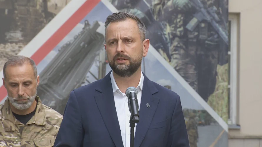 Mężczyzna w garniturze przemawia do mikrofonu na tle grafiki przedstawiającej żołnierzy i sprzęt wojskowy, obok niego stoi mężczyzna w mundurze wojskowym.