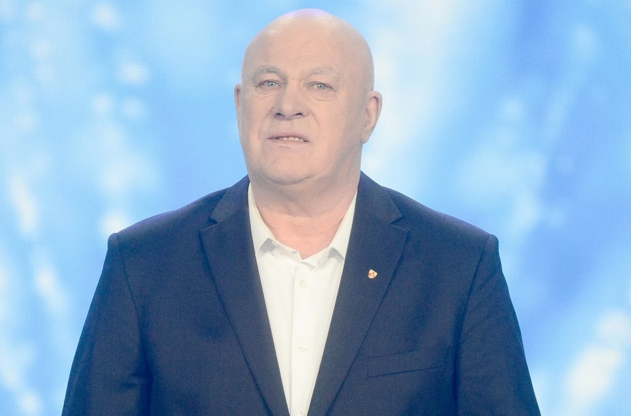 Ryszard Rynkowski usłyszał poważne zarzuty Starszy mężczyzna w garniturze i białej koszuli stoi na tle jednolitego, jasnoniebieskiego tła, ma lekko uśmiechniętą twarz i wykonuje spokojną, oficjalną pozę.